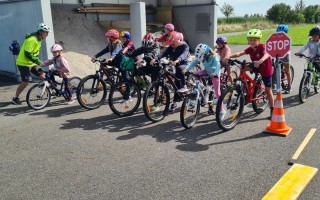 Kinder fahren auf asphaltierter Straße nebeneinander. Einige tragen helme. Vor ihnen eine gelbe Markierung. Im Hintergrund Grünflächen und ein Gebäude.
