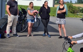 Drei Personen stehen auf einem Parkplatz vor einem Gebäude. Eine Person hält ein Baby. Im Hintergrund ein Fahrrad und ein weiteres Gebäude.