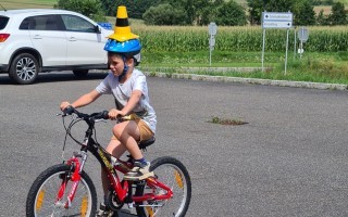 Ein Kind fährt Fahrrad auf einer Parkstraße. Es trägt einen gelben Helm und einen gelben Hut. Im Hintergrund ein Auto und ein Feld.