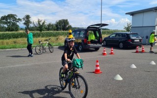 Ein Kind fährt Fahrrad auf einer asphaltierten Fläche mit aufgebauten Gelbmarkierungen und einem schwarzen Transporter im Hintergrund.