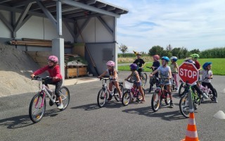 Kinder fahren Fahrräder auf einer asphaltierten Fläche. Im Hintergrund ein Schutzbau und eine Warnmarkierung.