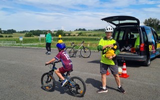 Ein Vater leitet einen Jungen beim Fahrradfahren auf einem Feldweg. Im Hintergrund steht ein gelber Kleinwagen mit gelben Warnwesten und Gelbwesten.