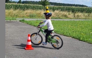 Ein Kind übt das Balancieren auf einem grünen Fahrrad auf einem asphaltierten Weg. Im Hintergrund befindet sich ein orangefarbener Konisch und ein Feld mit Mais.