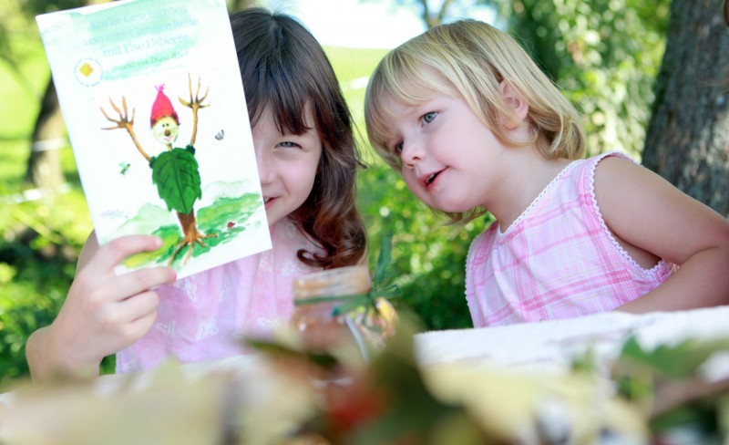 Zwei Kinder betrachten gemeinsam ein Bild mit einem kleinen Kobold im Grünen.