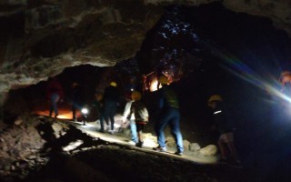 Gruppen von Personen bewegen sich auf einem schmalen Pfad in einem dunklen, felsigen Höhlengang. Scheinwerfer erzeugen Lichtreflexe.