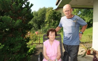 Zwei ältere Menschen sitzen im Freien. Eine Frau in rosa, eine ältere Mann in blau-weißem T-Shirt, sitzen in einem Garten.