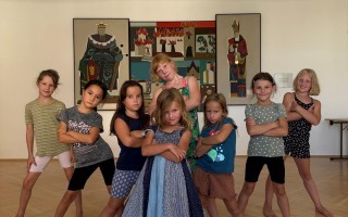 Mehrere Kinder, vermutlich im Alter von 8-12 Jahren, stehen in einer Gruppe und halten die Arme vor den Körper. Im Hintergrund hängt ein großes Kunstwerk.