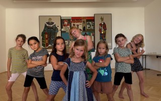 Mehrere Kinder stehen in einer Reihe und machen die Arme vor der Brust fest. Sie tragen verschiedene T-Shirts und Shorts. Im Hintergrund ein Wandbild.