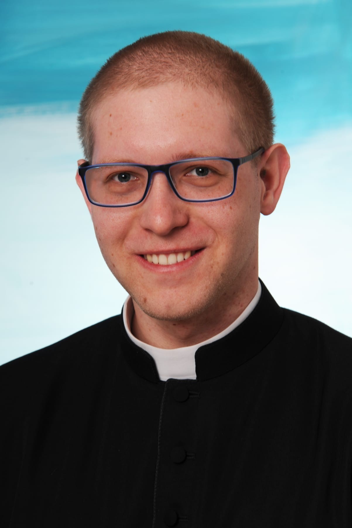 Mann mit hellem Haar, weißen Kragen und Brille. Er trägt eine schwarze Jacke mit weißem Kragen.