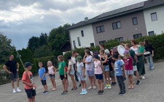 Kinder und Jugendliche in Trägersätzen in einer Gruppe auf einem Parkplatz vor einem Wohnhaus.