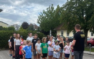 Kinder in Reihe, einige in bunten T-Shirts, vor einem Mann in Schwarz-T-Shirt, in einer belebten Umgebung mit Häusern im Hintergrund.