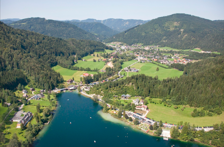 Flaches Tal mit See und Bebauung, umgeben von dichten Wäldern.