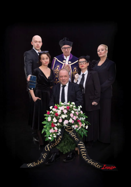 Schwarze Kleidung, fünf Personen im Gruppenbild, darunter ein Mann mit Krawatte und eine Frau mit Brille, sowie eine weiße Blumenwiese.