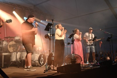 Bandmitglieder singen auf einer Bühne mit Schlagzeug und Gitarre im Hintergrund