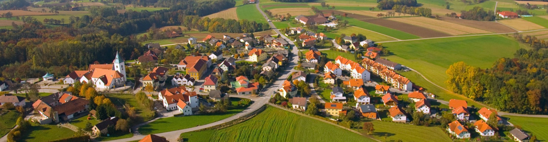 Erbauter Ort mit Häusern und Kirche, umgeben von grünen Feldern und Wiesen, aufgenommen aus der Vogelperspektive.