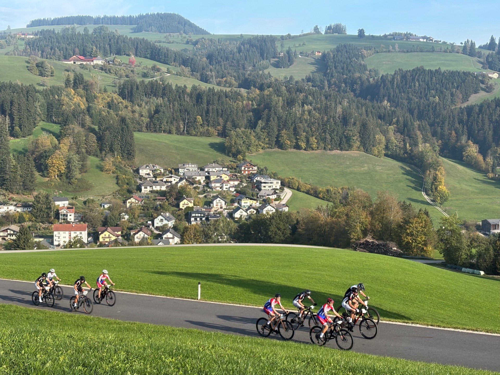 Radfahrer fahren eine Straße in der Steiermark mit hügeliger Landschaft und Häusern
