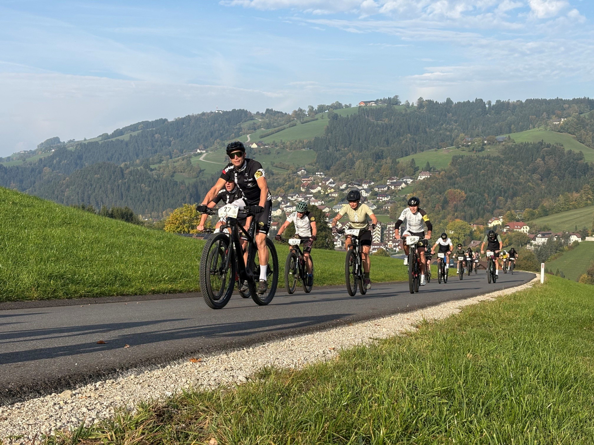 Mountainbiker fahren auf einer Straße in einer hügeligen Landschaft mit Häusern und Bäumen.